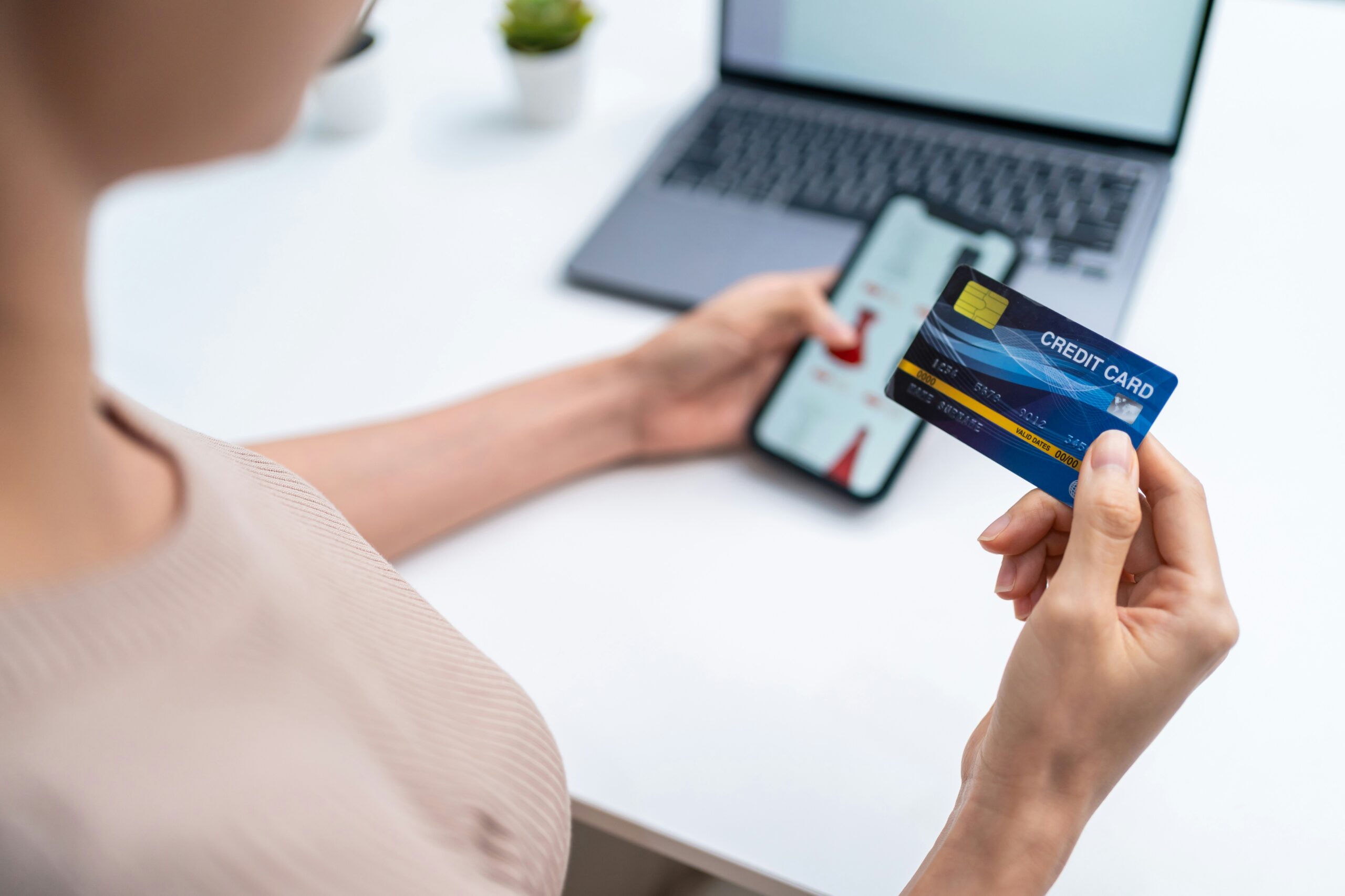 getty-images--bqawjDJWjo-unsplash Person holding a credit card in one hand and a mobile phone in the other, seated at a desk with a laptop and small potted plants in the background.