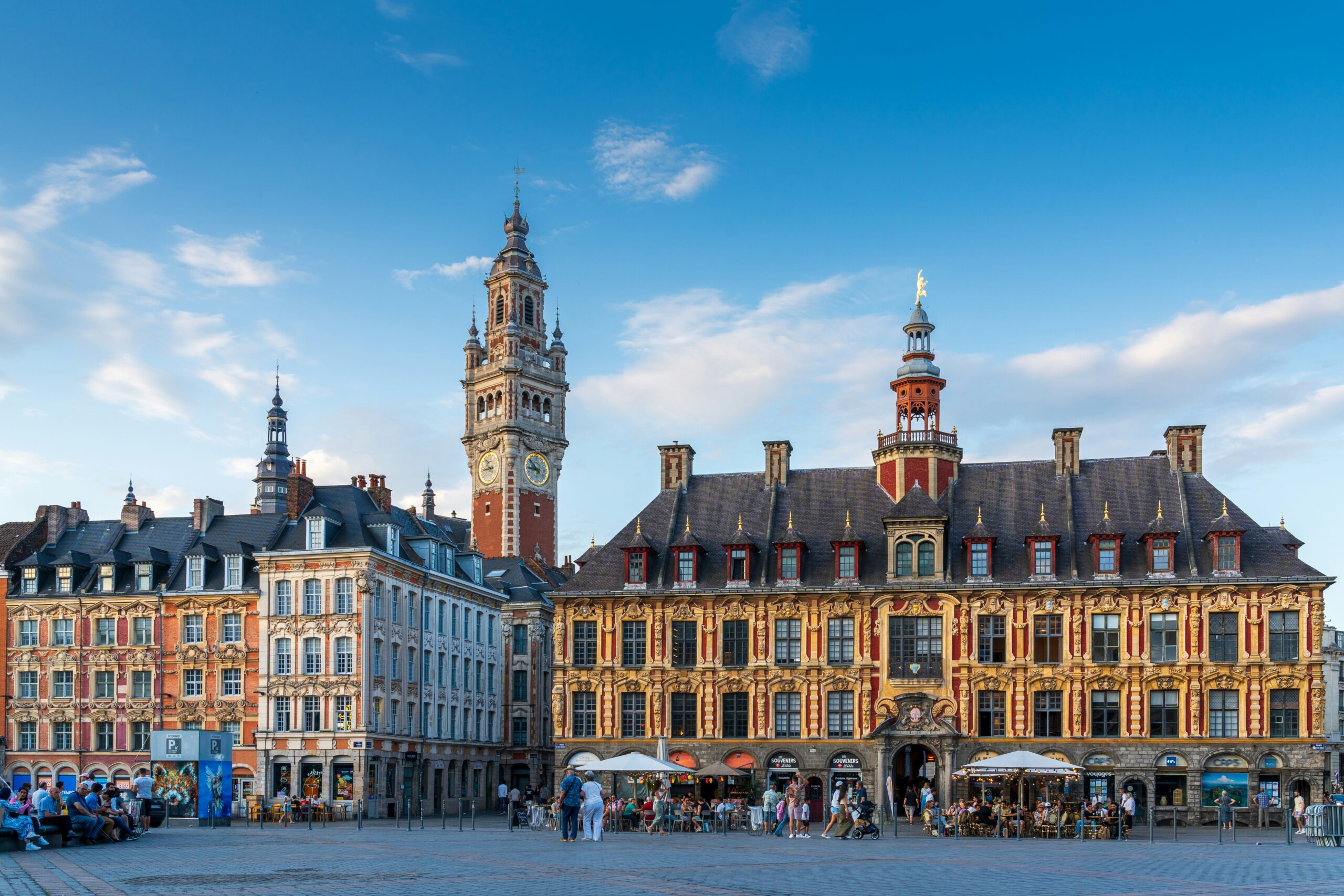 lille People walk and sit near cafés in a large square with ornate historic buildings and a clock tower under a clear blue sky.