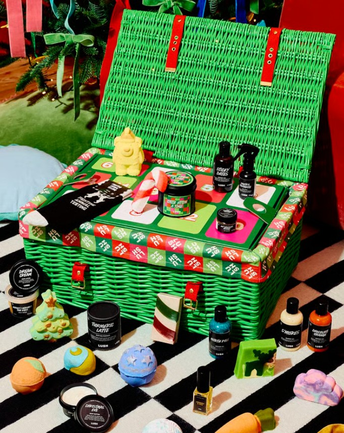 lush advent calendar 2026 A green wicker basket filled with assorted Lush bath and body products, surrounded by bath bombs and toiletries, sits open on a striped rug.