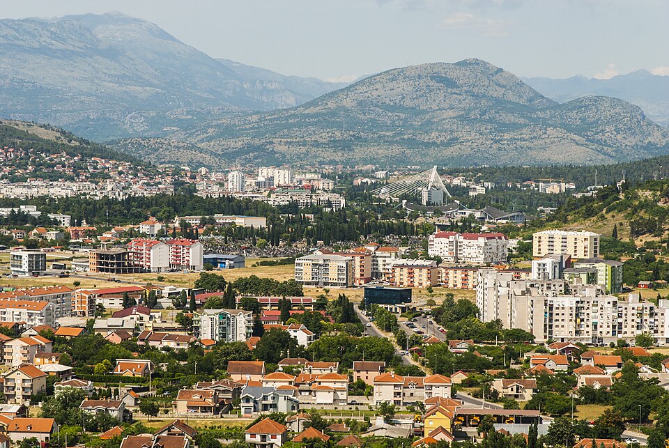 podgorica A cityscape featuring modern buildings with red-tiled roofs, tree-lined streets, a distant bridge, and mountains in the background under a clear sky.