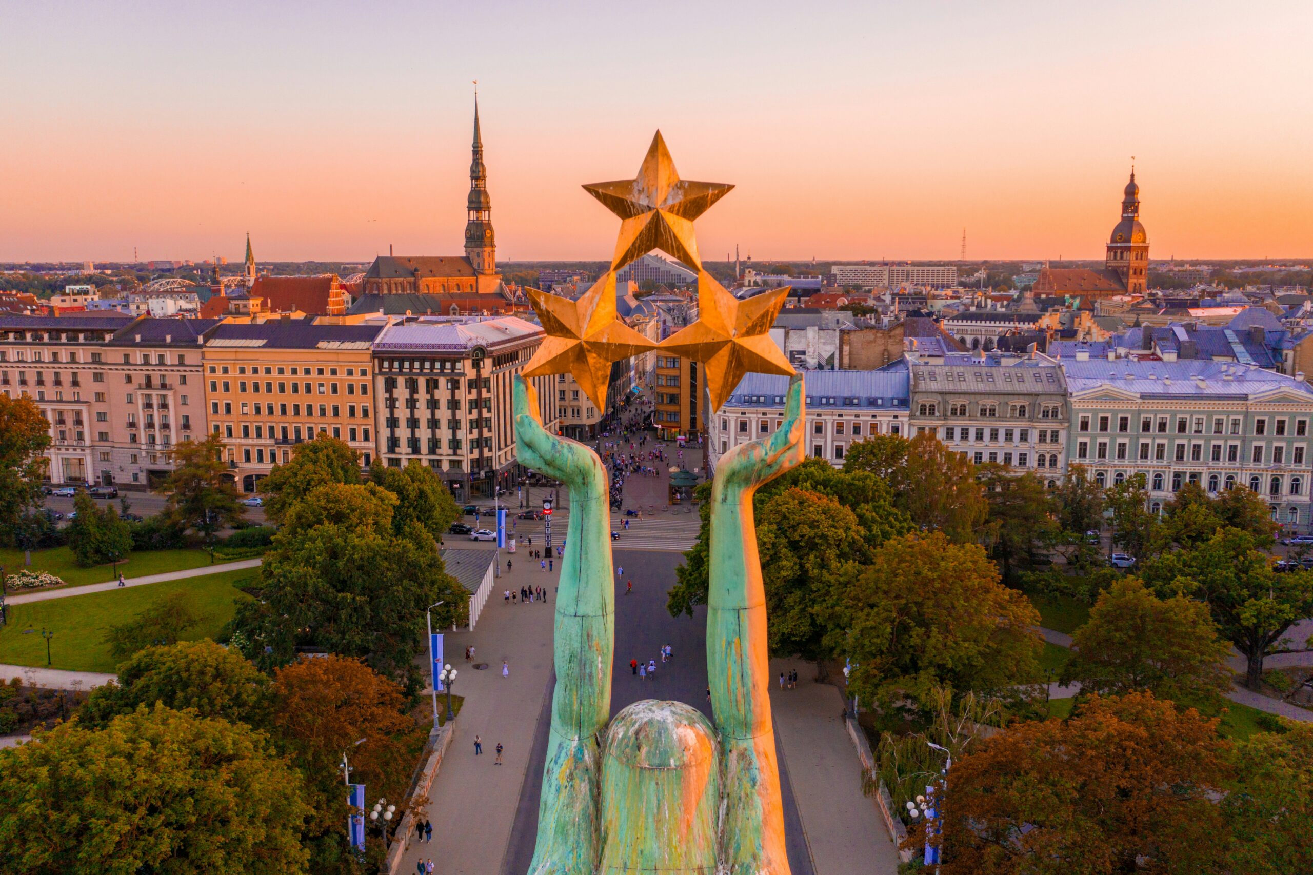 riga Aerial view of a large statue holding three stars, surrounded by trees and historic buildings at sunset in a cityscape.