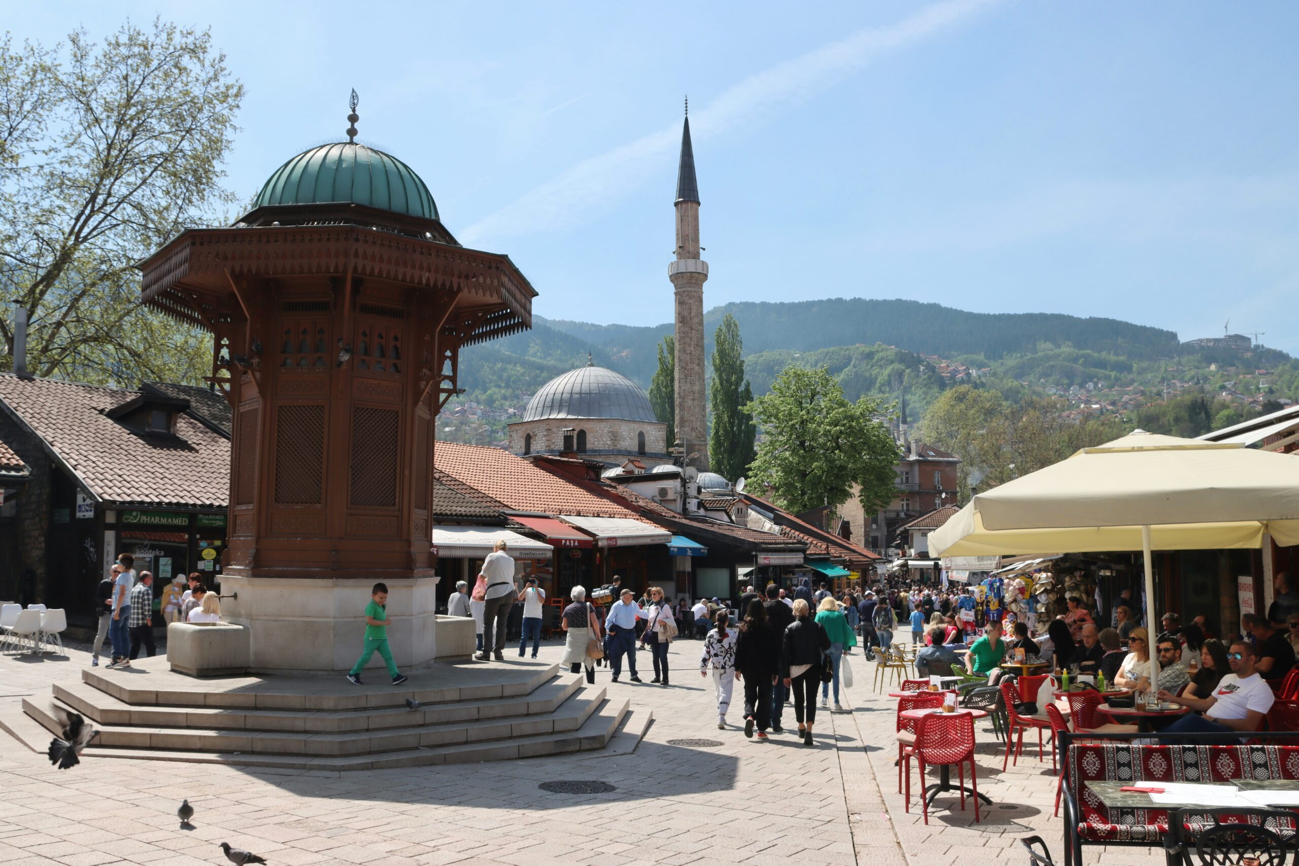 sarajevo Pedestrians walk through a lively outdoor square with a wooden fountain, market stalls, cafés, and a mosque with a tall minaret in the background.