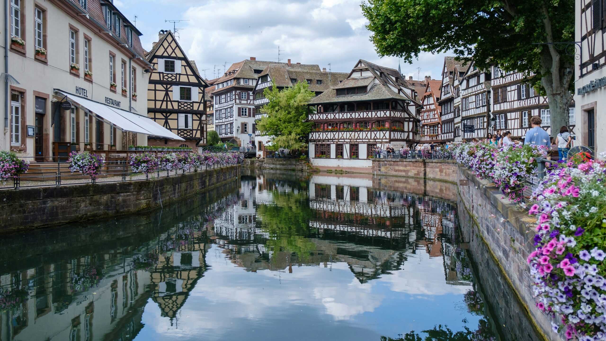 strasbourg A canal runs between half-timbered buildings with flower boxes on both sides, reflecting in the water under a partly cloudy sky.