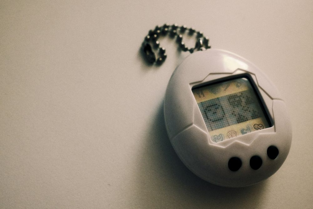 tamagotchi A close-up of a white digital pet device with three black buttons and a chain, displaying pixelated graphics on its screen.