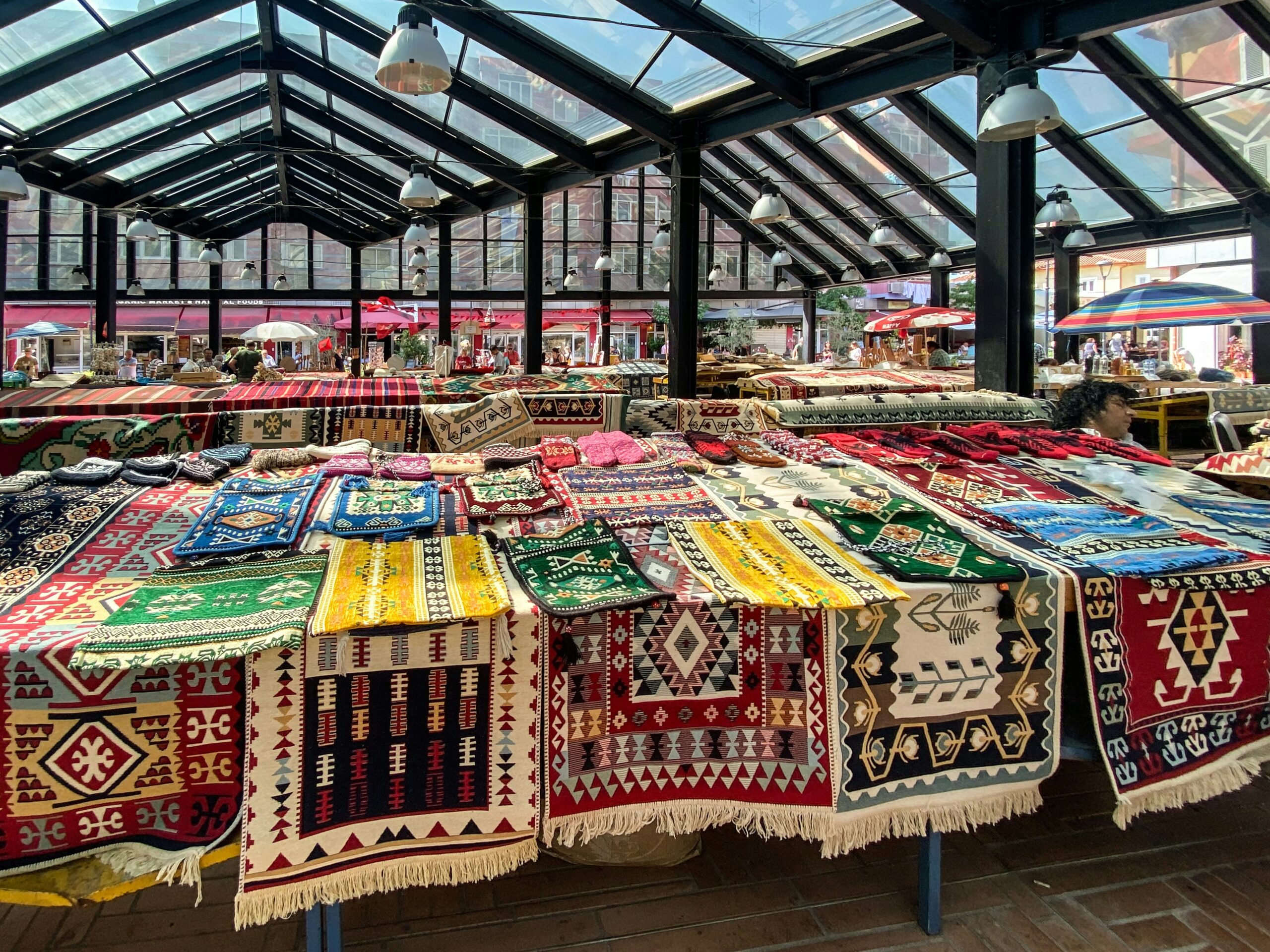 tirana Market stall under a glass roof displays colourful patterned rugs and textiles arranged on tables, with more market activity visible in the background.
