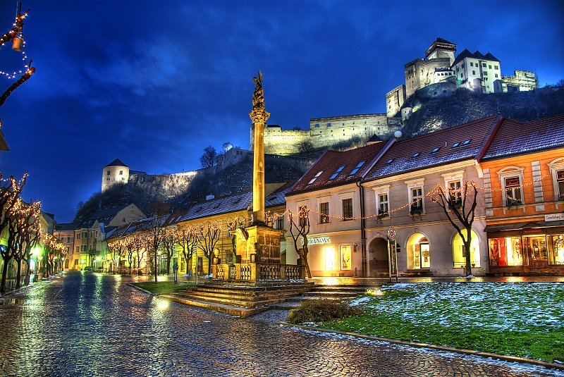 trencin slovakia A cobblestone street with small shops, a central monument, and a large hilltop castle illuminated at twilight.