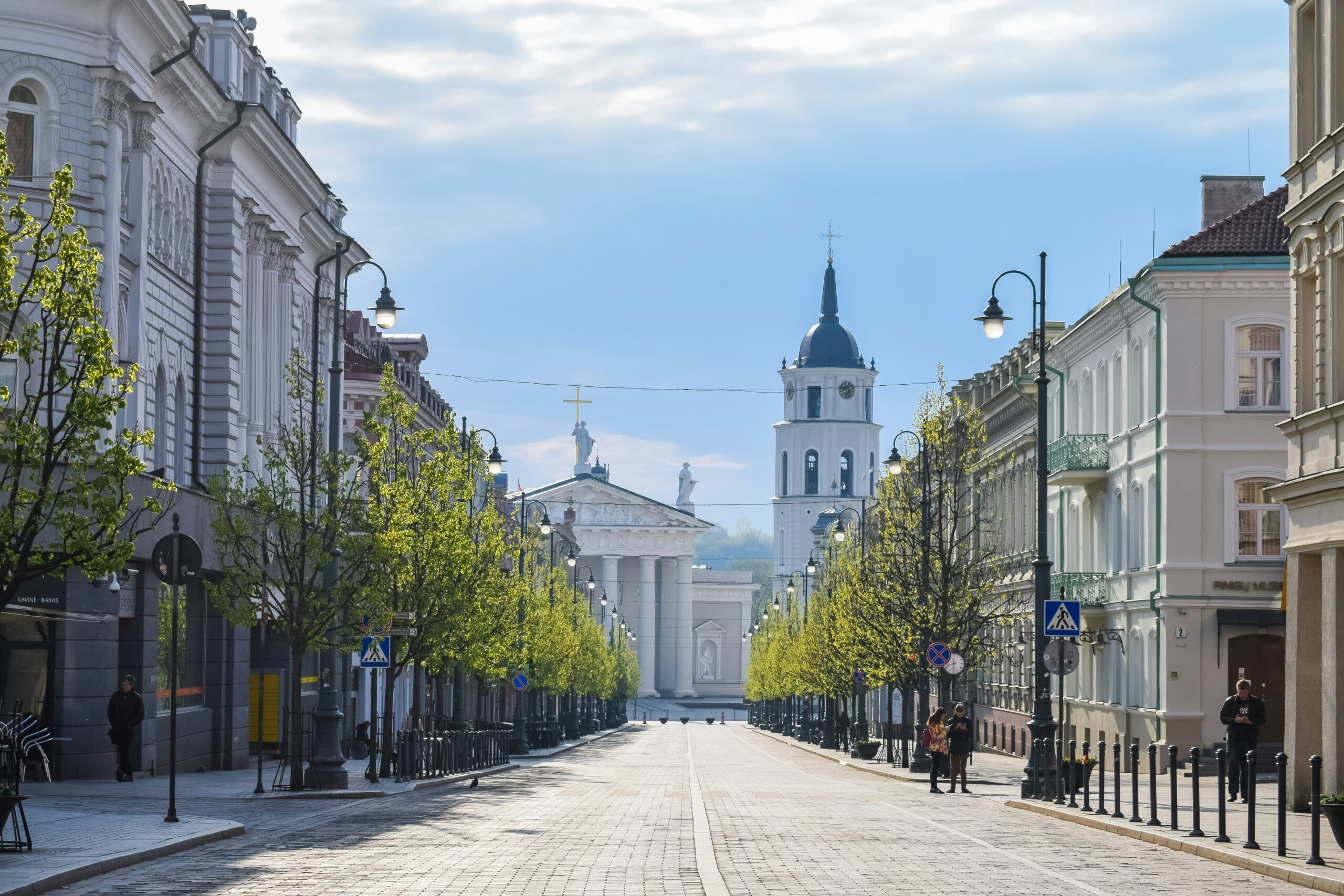 vilnius A wide cobbled street lined with trees and historic buildings leads to a white church with a tall bell tower under a cloudy sky.