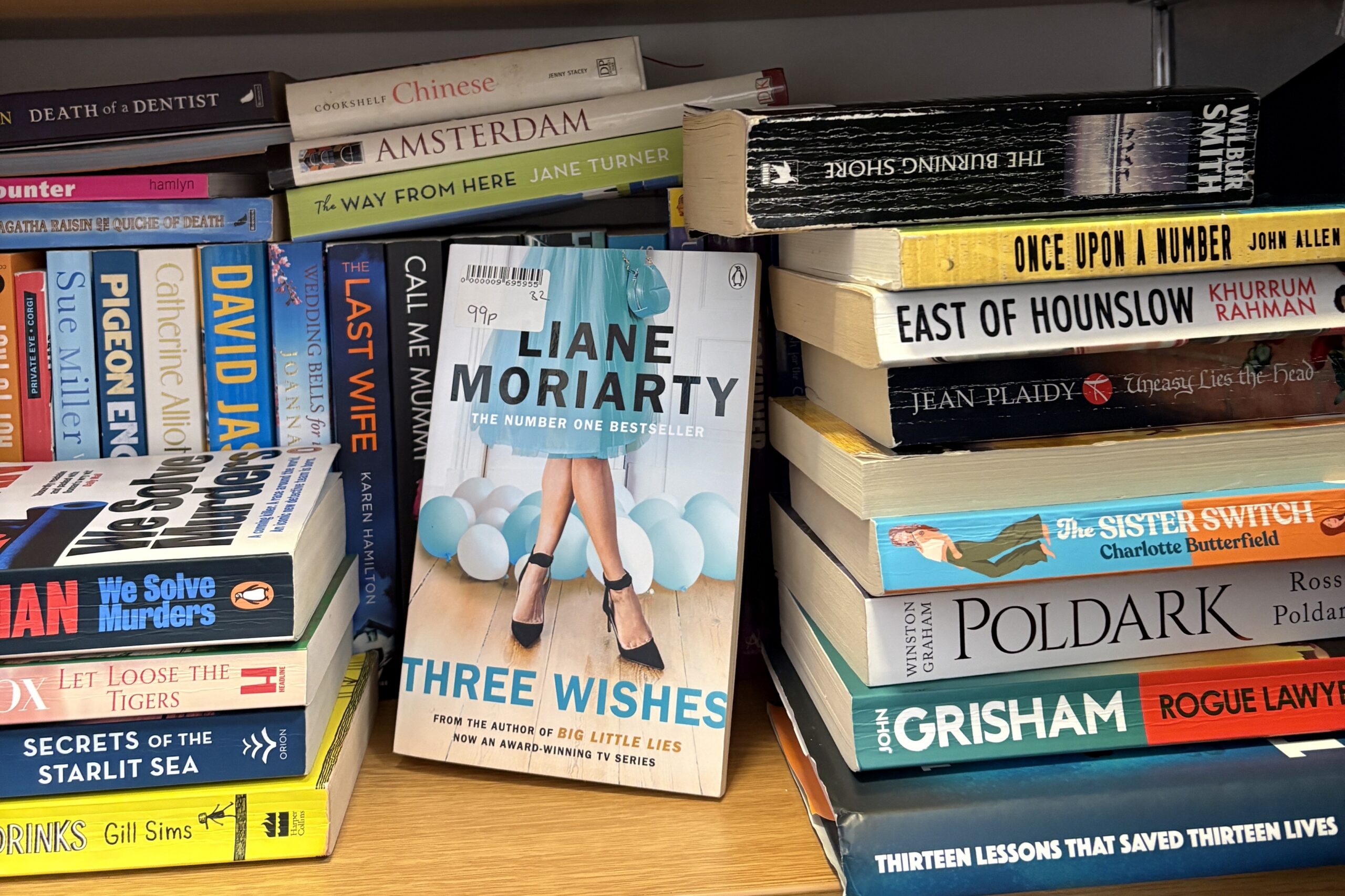 charity shop books A bookcase displays various novels, including “Three Wishes” by Liane Moriarty in the centre, surrounded by other fiction and crime books stacked horizontally and vertically.