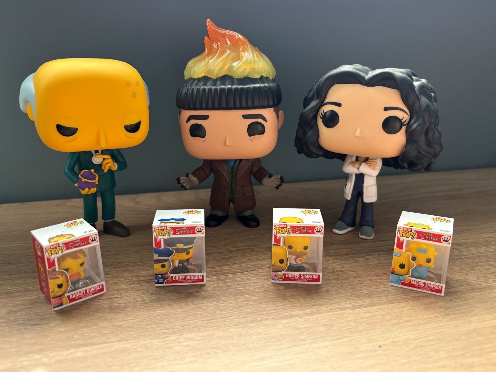Three Funko Pop figures stand behind four miniature boxed Funko Pop figures, all displayed on a wooden surface against a plain blue-grey background.