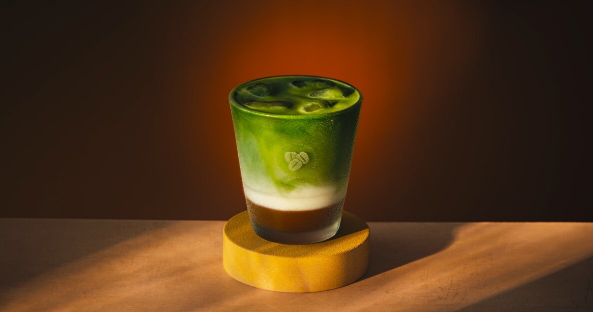 maple hazel iced matcha latte costa coffee autumn 2026 A glass of iced matcha latte with distinct green, white, and brown layers sits on a round wooden coaster, against a dark brown background.