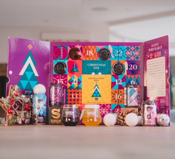 siren craft brew beer advent calendar 2026 Colourful advent calendar box with numbered doors, surrounded by tinned drinks, decorations, pine cones, and Christmas-themed items, arranged on a wooden surface.