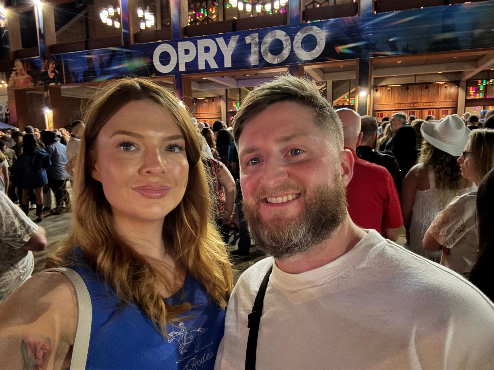 Two people pose for a selfie at night in front of a crowd outside a building with a sign reading “OPRY 100”.