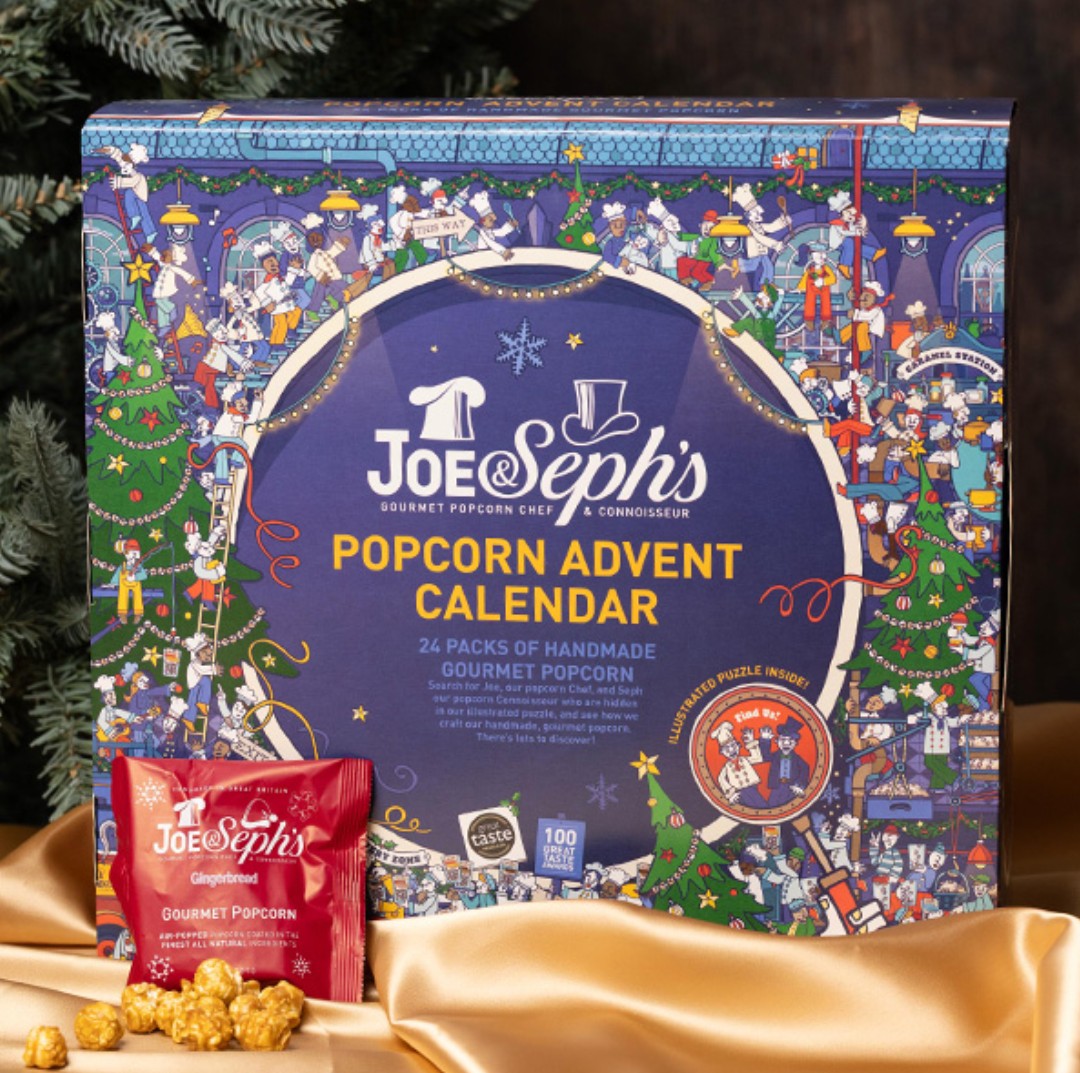 joe and sephs gourmet popcorn advent calendar 2026 Joe & Seph’s Popcorn Advent Calendar box with festive illustrations, a small red popcorn packet, and pieces of caramel popcorn displayed in front on a gold cloth near a tree.