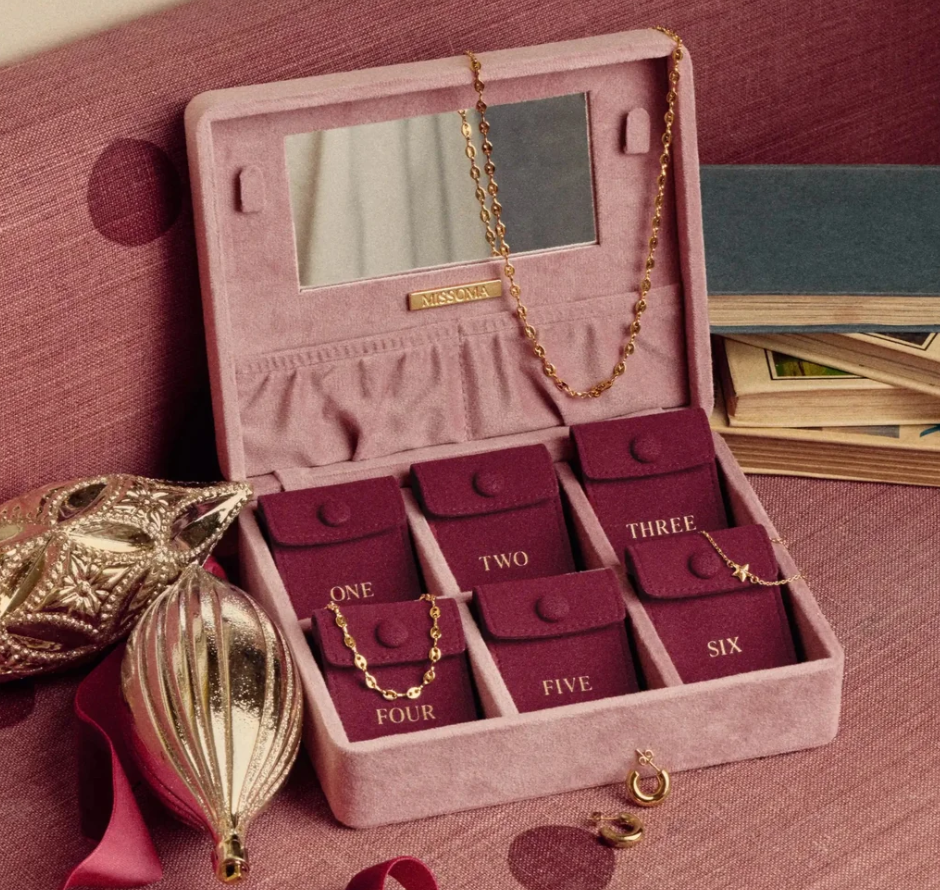 missoma gold advent calendar 2026.jpg A pink jewellery box with six compartments labelled one to six, holding gold jewellery, sits on a pink surface beside holiday ornaments, a ribbon, and a stack of books.