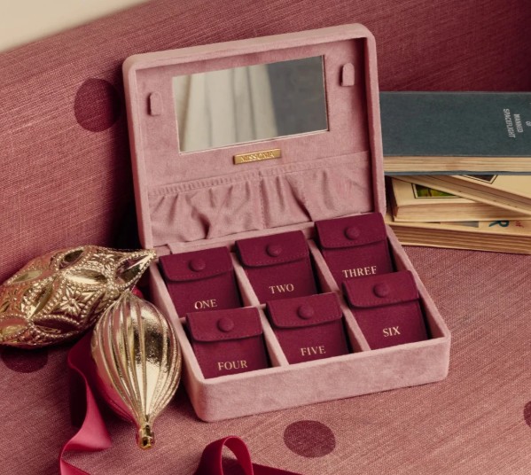 A pink velvet box with a mirror contains six labelled pouches numbered one to six, next to a book and two decorative ornaments on a pink surface.