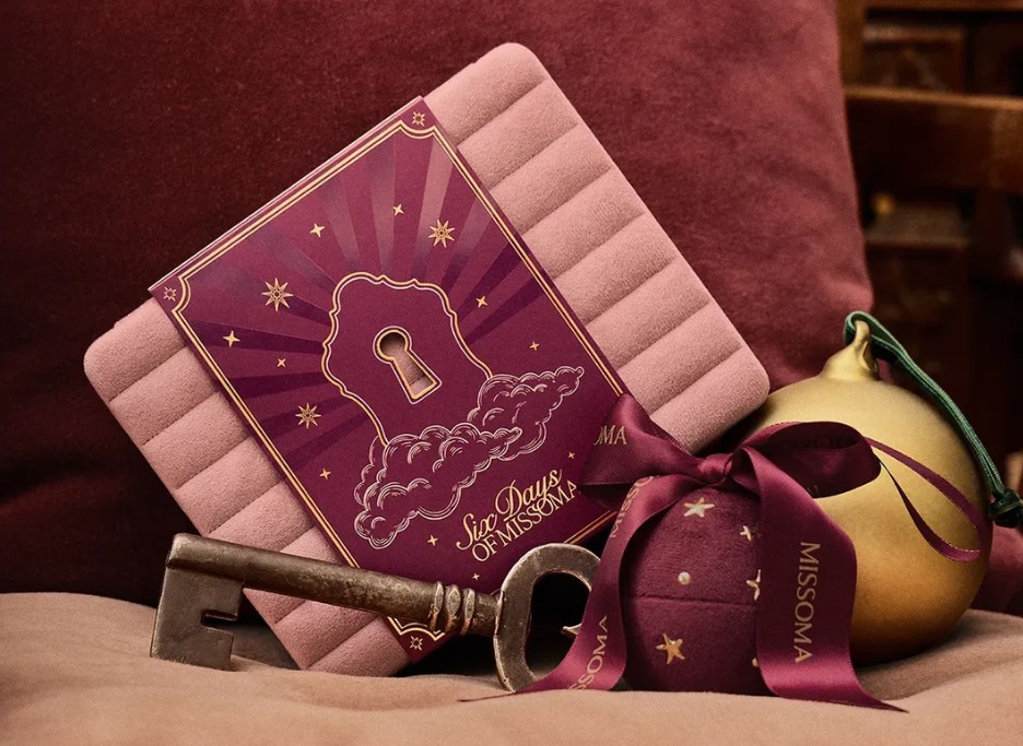 missoma jewellery advent calendars 2026 A vintage key, a gold ornament, and a maroon box with “Six Days of Missoma” and a ribbon sit on a soft pink cushion.