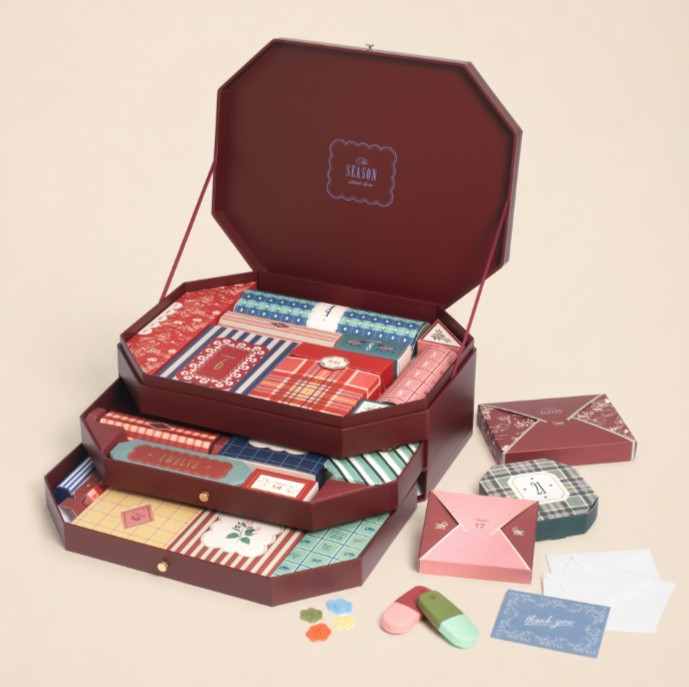 papier 24 day advent calendar A three-tiered maroon gift box is open, displaying various patterned stationery items, envelopes, cards, and small colourful objects arranged neatly inside and around the box.