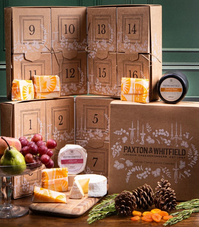paxton and whitfield cheese advent Advent calendar boxes labelled 1–15, Paxton & Whitfield cheeses, fruits, nuts, and herbs arranged on a table in front of a green panelled wall.