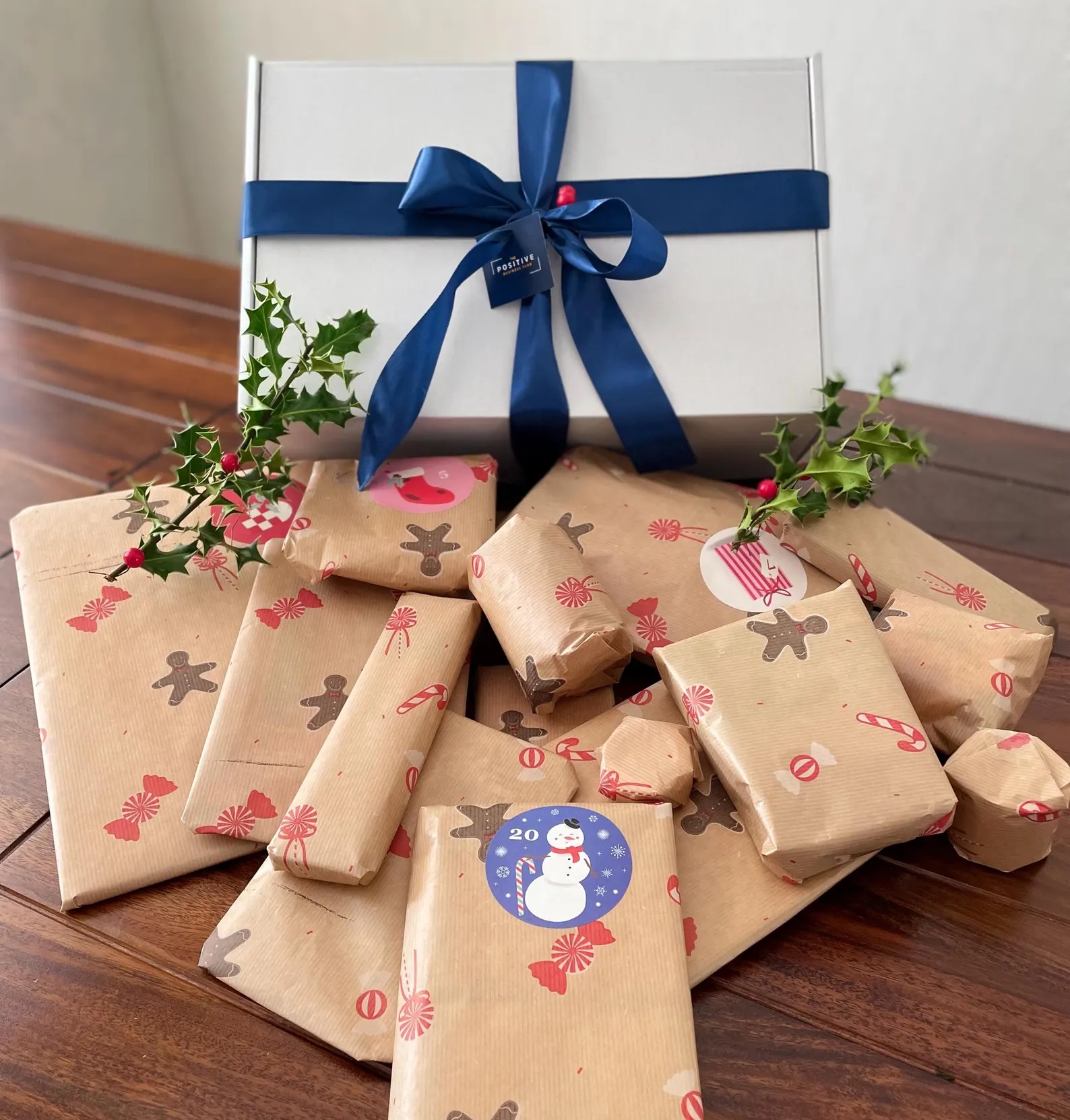positive business club advent calendar A group of wrapped presents in brown paper with gingerbread and festive designs, plus a white box tied with a blue ribbon, on a wooden table with sprigs of holly.