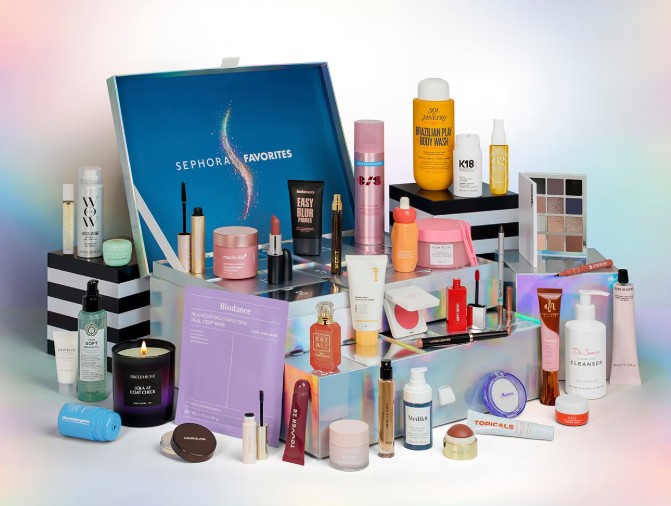 sephora advent calendar 2026 .png A large assortment of beauty and skincare products, including creams, serums, make-up, and candles, is arranged in front of an open Sephora Favourites box on a reflective surface.