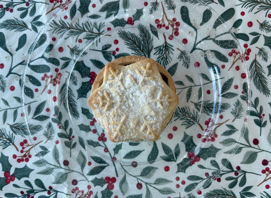A mince pie topped with a snowflake-shaped pastry lid dusted with icing sugar on a glass plate over a holiday-themed tablecloth.