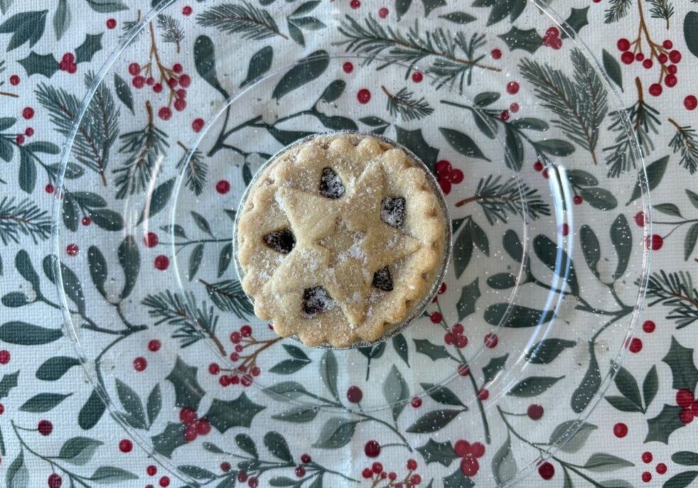 A single mince pie with a star-shaped cut-out on a clear plate over a holiday-themed tablecloth.