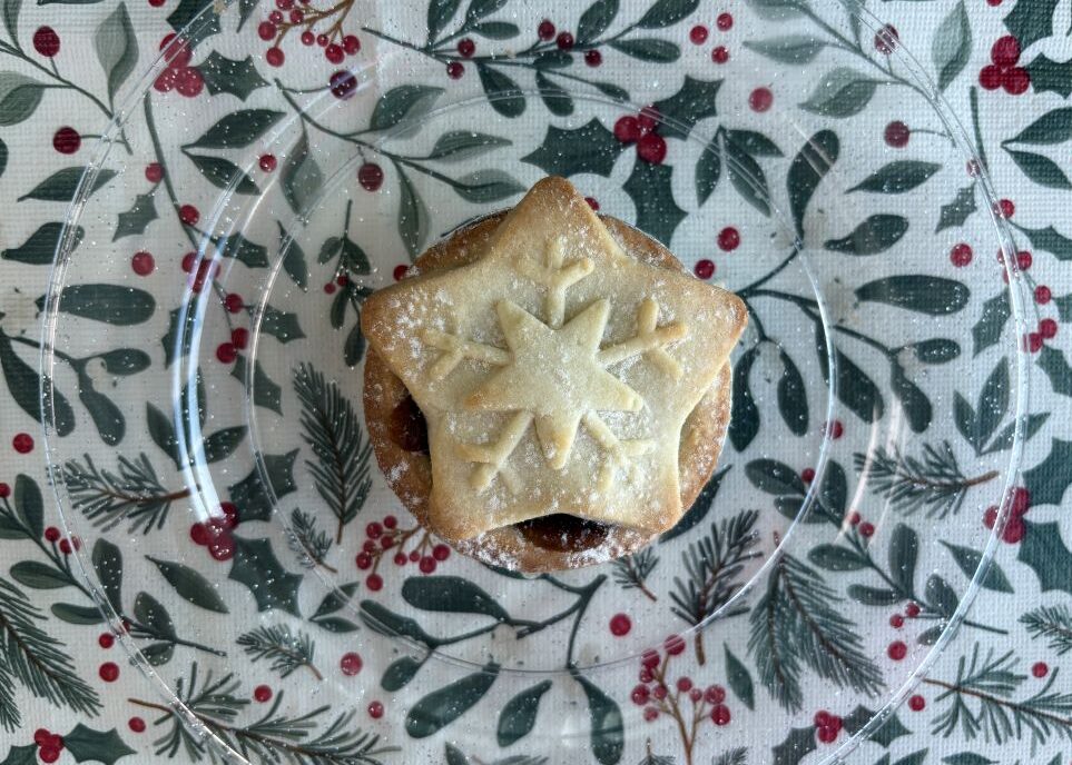 A mince pie topped with a pastry snowflake sits on a clear plate over a holiday-themed tablecloth.