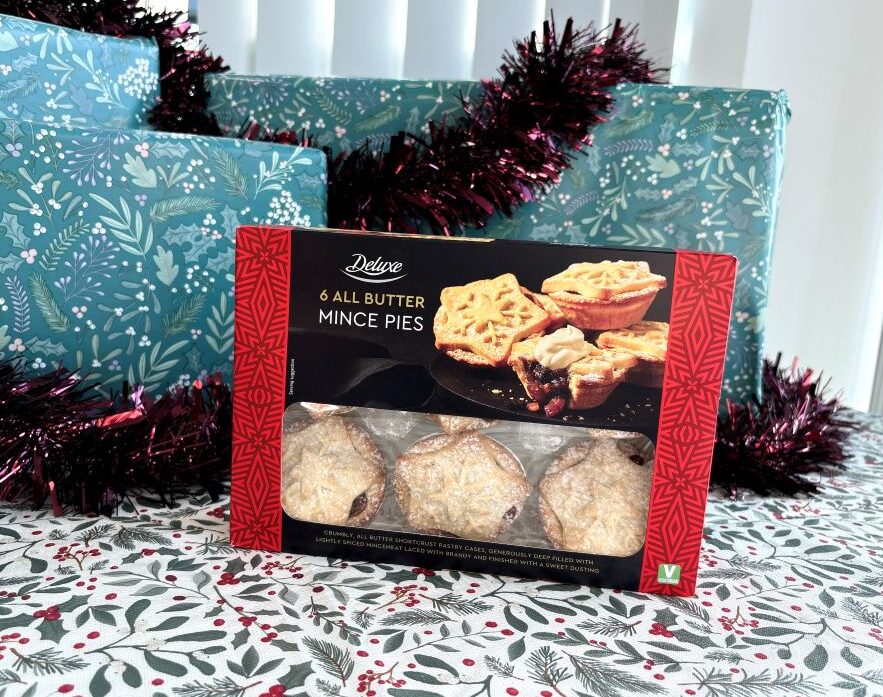 A box of six Deluxe all-butter mince pies positioned in front of wrapped Christmas presents and tinsel.