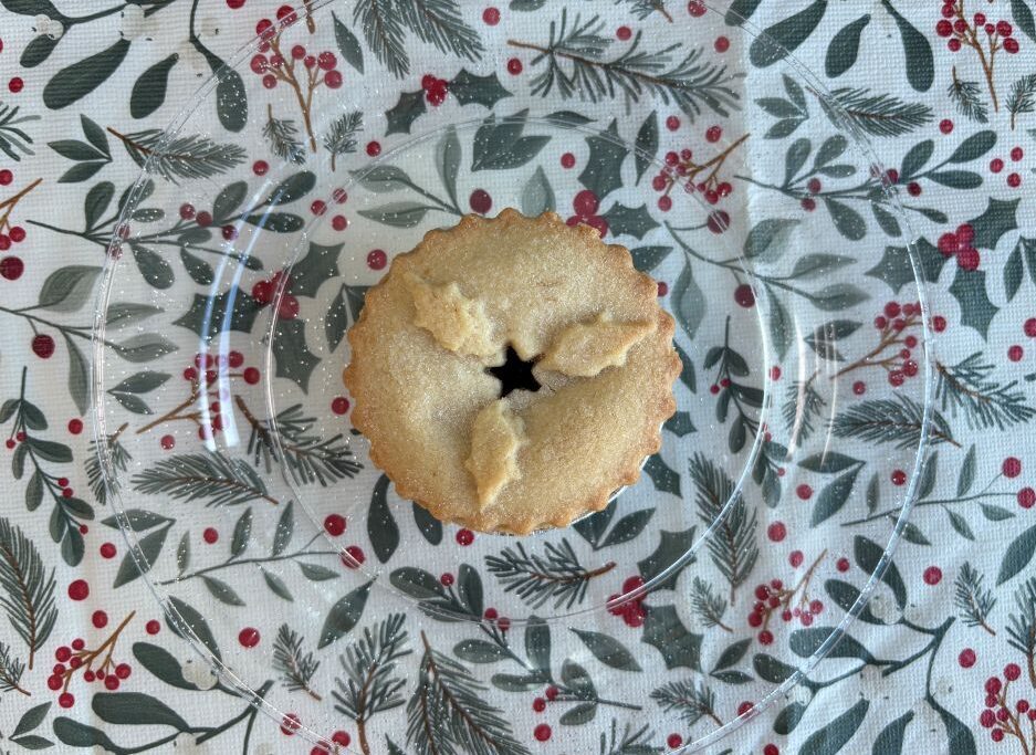 A single mince pie with a star-shaped cut-out on a clear glass plate over a holiday-themed tablecloth.