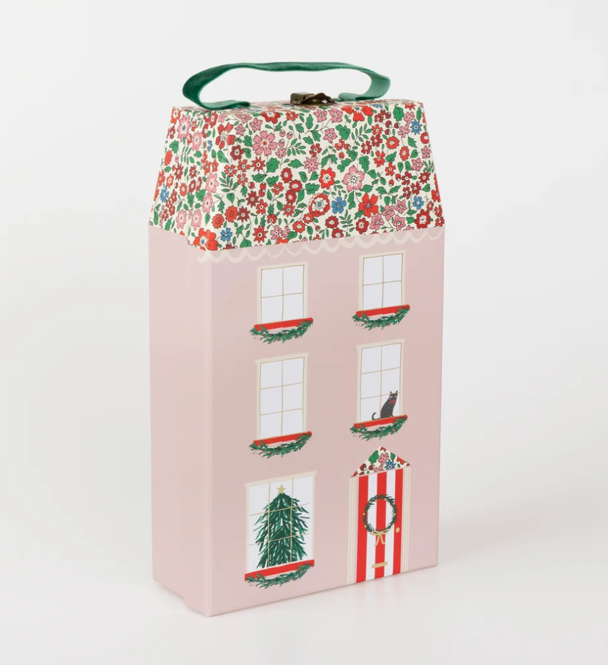 meri meri liberty advent calendar A pink, house-shaped gift box with a floral roof, a green ribbon handle, and illustrations of windows, a door, and holiday decorations.