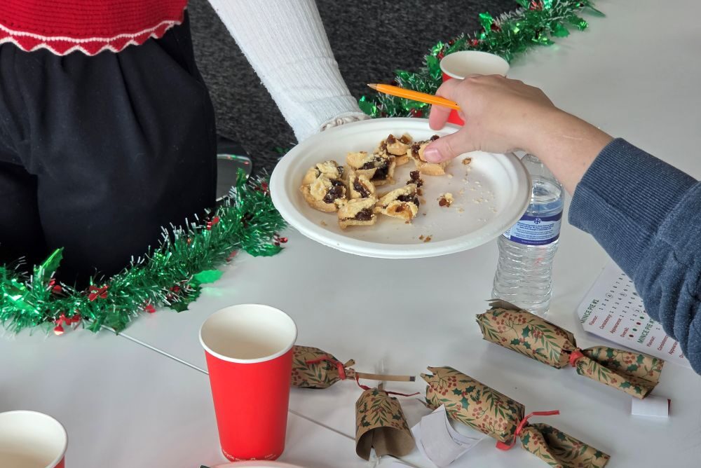 A hand holding a pencil reaches for a plate of mince pie pieces on a table decorated with Christmas crackers and tinsel.