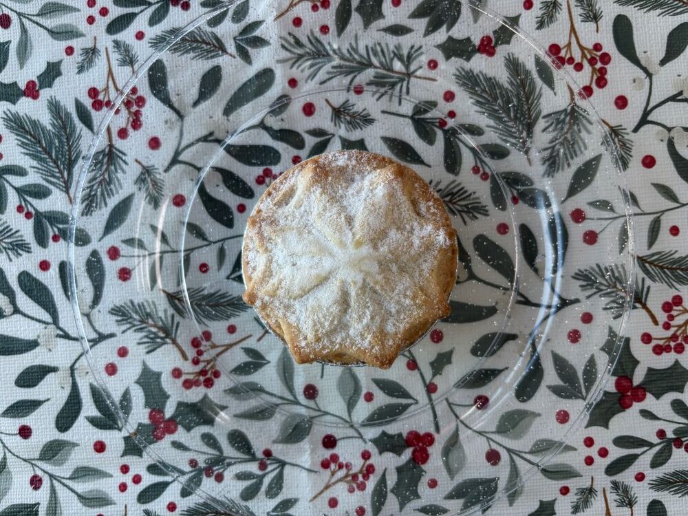 A star-shaped mince pie dusted with icing sugar on a clear plate over a holiday-themed tablecloth.