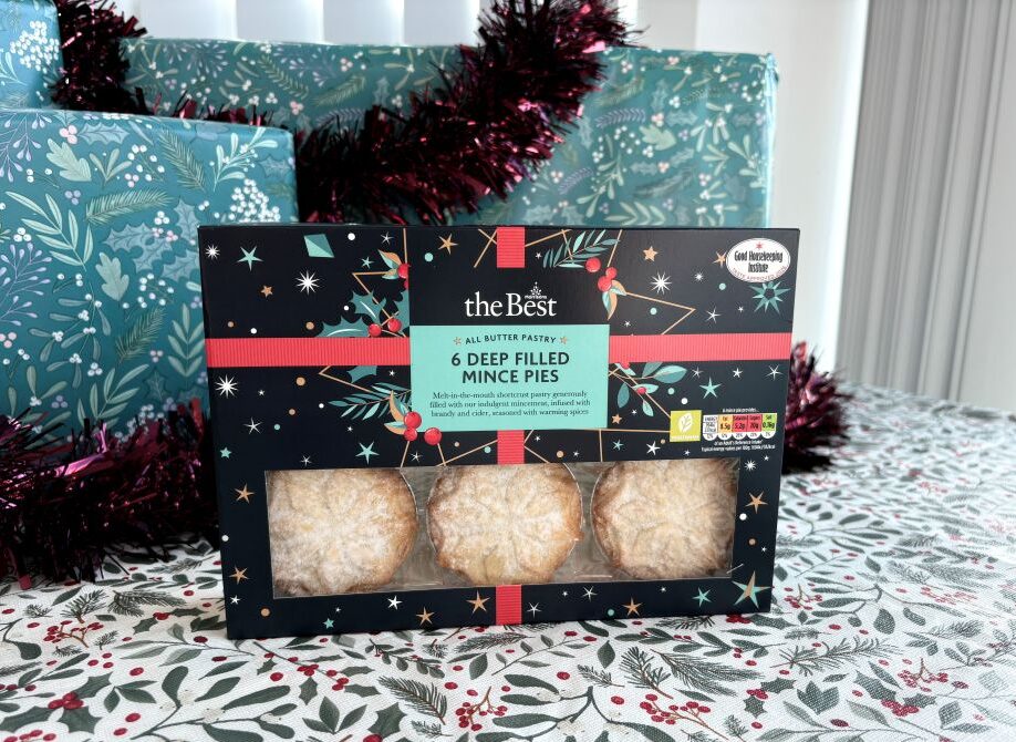 Box of six deep-filled mince pies decorated with a holiday pattern, placed on a festive tablecloth with wrapped presents in the background.