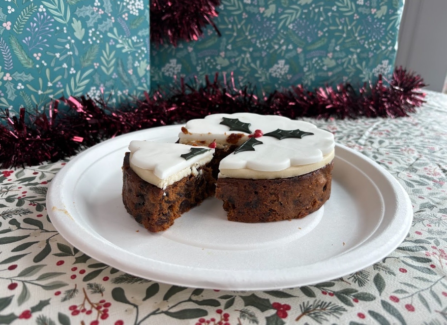 m&s christmas cake 2026 A sliced fruit cake topped with white icing and holly decorations on a paper plate, in front of holiday-themed gift wrap and tinsel.