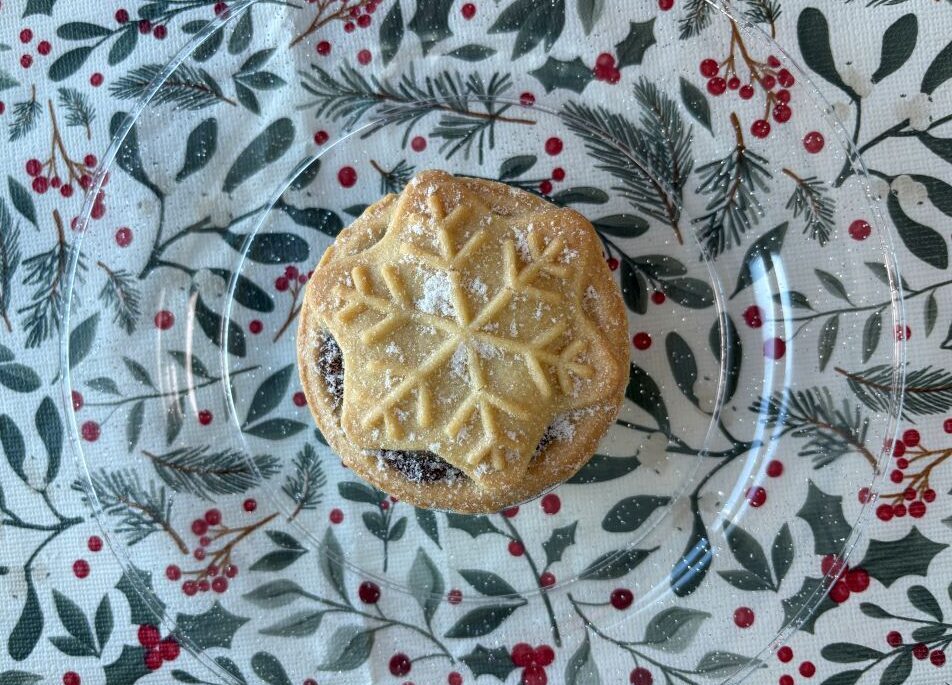 A fruit mince pie with a snowflake-shaped pastry top on a clear glass plate over a holiday-themed tablecloth.