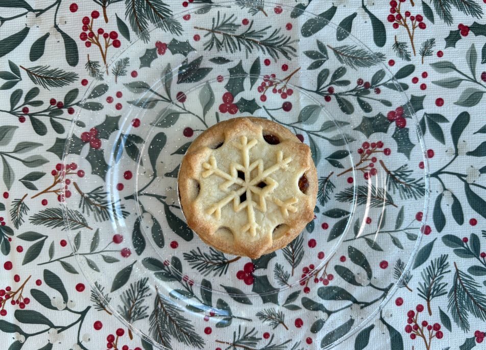 A single mince pie with a snowflake-shaped pastry cut-out, centred on a clear glass plate over a holiday-themed tablecloth.